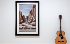A guitar stands against a wall with a large painting.